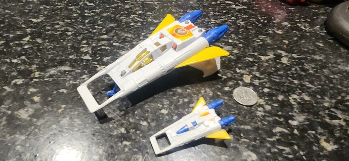 Same two white toy spaceships with blue engine nozzles on a speckled black counter top, 25c coin for unhelpful size comparison. But now they have yellow wings extended for atmospheric flight..