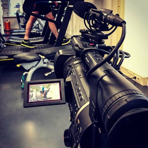 A videocamera is filming someone on a treadmill. The electronic viewvinder shows the whole person (very small) and the complete picture only the treadmill and some legs. The focus is on the Panasonic AG130 videocamera with cables and shotgun microphone.