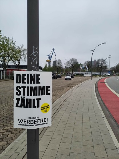 An einer recht neuen Straße, an einem Laternenmast: A1-Plakat mit "Deine Stimme zählt. Hamburg Werbefrei - unterschreib bis zum 13. Mai"