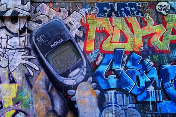 The image features a colorful graffiti mural with a prominent depiction of a classic Nokia mobile phone. The background is filled with various vibrant graffiti tags and designs, showcasing a mix of colors and artistic styles. The phone appears to be held by a hand, blending with the urban art scene.