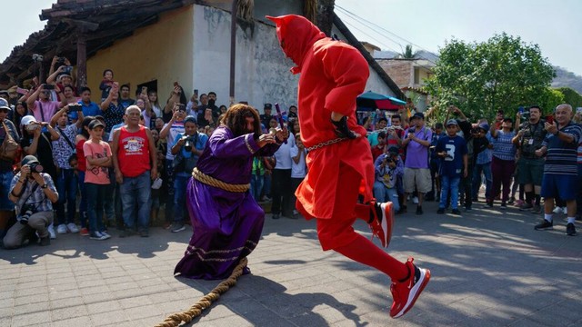 Ator fantaseado de Jesus segurando um crucifixo e apontando para o outro ator, fantasiado de Diabo.
