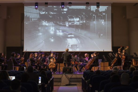 L’orchestra d’archi Young del Conservatorio di Foggia esegue QFD di Nicola Monopoli. Francesco Greco suona il violoncello e Andrea Palmacci dirige, sullo sfondo il live visuals di Simona Dichio, le strade della città di Copenaghen in bianco e nero.