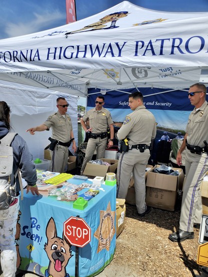 Boxes of bicycle helmets are stacked behind four California Highway Patrol uniformed officers at their SOC booth. Reflective straps, stickers, brochures, and inexpensive lights are displayed on the table before them.
