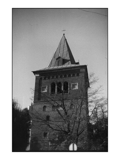 The image depicts a tall brick tower with a pointed roof, topped by a cross, suggesting it is part of a church or religious building. The tower features arched windows on the upper levels and rectangular windows on the lower levels, with a series of small, square windows near the base. The structure is surrounded by bare trees, indicating a possibly cold season. The photograph is in black and white, giving it a historical or vintage appearance. The sky is clear, and the overall composition emphasizes the tower's height and architectural details.