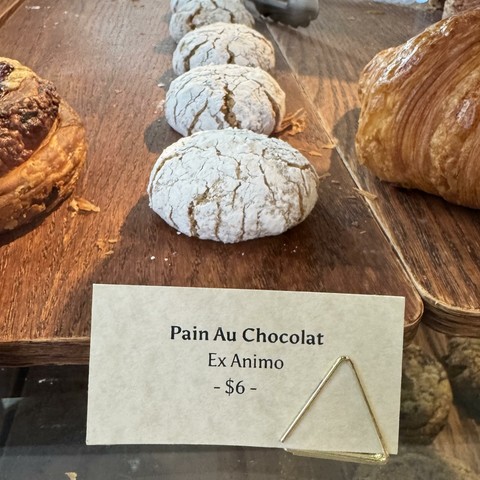 Photo of a row of flour-covered Amaretti on a wooden board. A paper sign in front of the first one advertises Pains Au Chocolate.