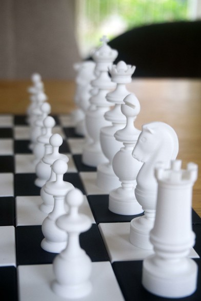 Closeup of a 3D printed chess set and chess board on a tabletop. The photo focuses on the white pieces, photographed from a side view angle.