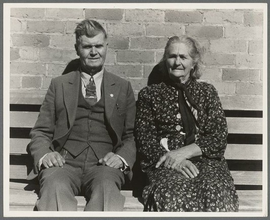 The image is a black and white photograph depicting an elderly couple sitting on a bench against a brick wall. The man, wearing a suit with suspenders and a tie, has medals pinned to his chest which suggest some form of recognition or honor. He also wears glasses around the neck.
Beside him sits a woman dressed in a floral-patterned dress with a dark shawl draped over her shoulders. She appears older than the man but cannot be identified through gender as she is wearing more traditionally feminine clothing for that era, and has no visible jewelry apart from rings on both hands. They are sitting close together; their proximity indicates familiarity or relationship.
The background consists of only plain brick walls which provides contrast against them making it clear they're likely in an outdoor setting such as a park bench typically found outside public buildings like schools or churches during that period given the couple's attire suggesting formality and importance to occasion.