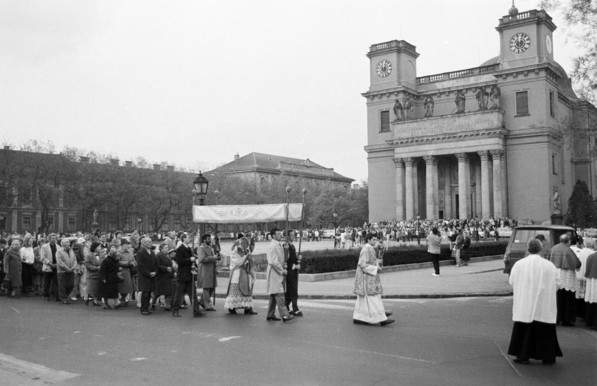 A black and white photo showing a large, imposing church with square towers on either side and Romanesque pillars in the front. There is a large square in front of the church where people are milling about. The steps of the church are also crowded with people.  A large crowd is also in a procession around the church.