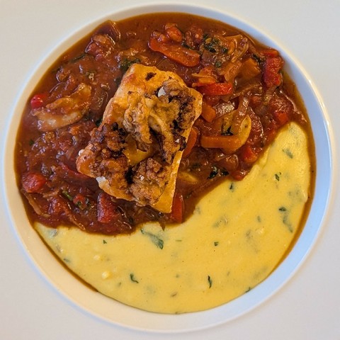 White bowl half filled with chunky red stew topped with cod and crisp tentacles, half filled with bright yellow herb-flecked polenta