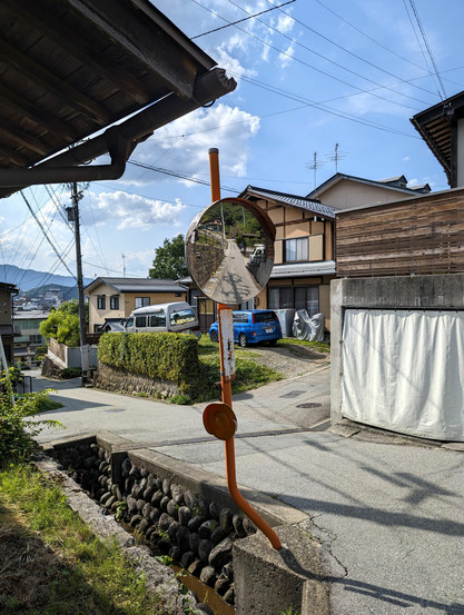 A road mirror with a curved pole to avoid being hit by vehicles taking the turn. The mirror is above a small water canal and reflects the road all the way up until a forested area. The background is a sunny clear summer sky and a Japanese residential area with houses and cars.