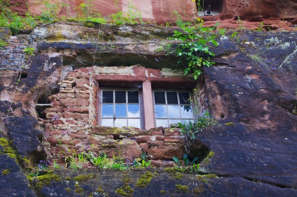 Eine Wand aus teilweise schwarz verfärbtem roten Sandstein. In den Stein gehauen ist eine Nische, zurückversetzt von der nHauptwand und grob mit alten Ziegeln und Steinen ummauert. Darin ein Doppelfenster mit jeweils 9 Teilen, die jeweils obere Reihe sind netze, die 2 Reihen darunter Scheiben. In denen spiegelt sich der Himmel mit Wolken. Der äußere Sandstein ist leicht bewachsen mit diversen kleinen Pflanzen. Ein paar mit zwarten Blüten in gelb und lila.