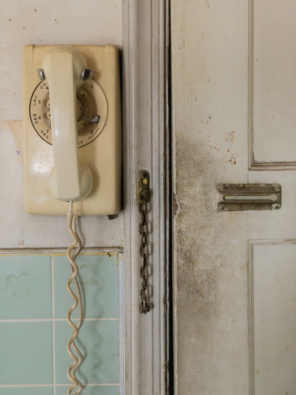 An old rotary telephone with a long, coiled cord, mounted on a dirty wall next to a dirty door with an unlocked chain latch.