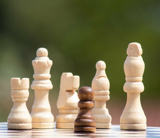 Een opstelling op een schaakbord: dicht bij de lens van de camera staat één zwarte pion. Om hem heen staan onscherp vijf witte schaakstukken: een toren, een koningin, een paard, een loper en een koning.