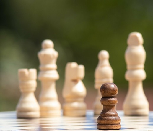 Een opstelling op een schaakbord: dicht bij de lens van de camera staat één zwarte pion. Om hem heen staan onscherp vijf witte schaakstukken: een toren, een koningin, een paard, een loper en een koning.