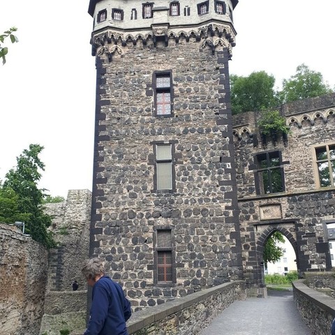 Das Bild zeigt eine mittelalterliche Steinburg mit einem hohen, zylindrischen Turm, der aus dunklen und hellen Steinen gebaut ist. Der Turm hat mehrere Fenster mit schmiedeeisernen Gittern und ist mit gotischen Verzierungen an der Spitze versehen. Neben dem Turm ist ein Torbogen zu sehen, der ebenfalls aus Stein gefertigt ist und mit einem steinernen Wappen verziert ist. Der Weg vor dem Torbogen führt zu einem gepflasterten Bereich, auf dem ein Person mit grauem Haar und einem blauen Pullover spazieren geht. Im Hintergrund sind Bäume und ein Teil eines modernen Gebäudes zu erkennen. Die Burg ist von einer Mauer umgeben, die teilweise von Pflanzen überwuchert ist.

Bereitgestellt von @altbot, privat und lokal generiert mit Ovis2-8B
