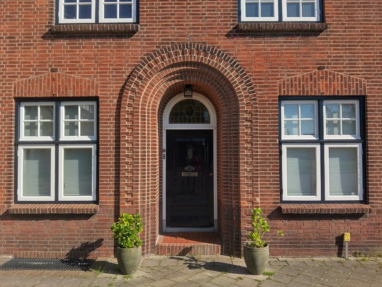 Foto van een facade uit bakstenen met een deur onder een halfronde poort met twee ramen aan beide kanten | Photo of a brick facade with a door under an arched gate and two windows on both sides