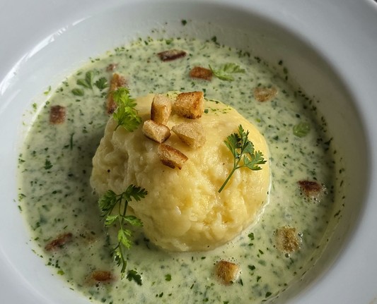 ein großer runder Kartoffelkloss in einem Teller, umgossen mit hellgrüner Suppe, in der Kräuterfitzelchen zu sehen sind, mit braunen Croutons und ein paar Kerbelblättchen ausdekoriert