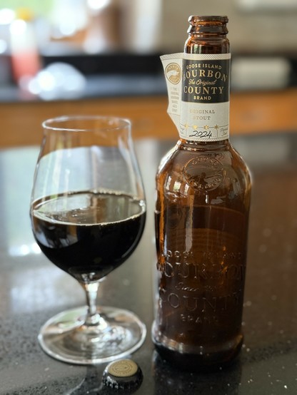 A snifter glass half full of dark beer next to a tall brown bottle with a white neck label with a goose outlined on gold