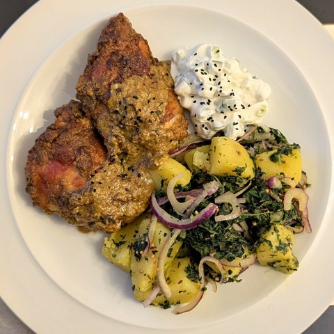 Two crisp chicken thighs with a little pale yellow brown sauce, pile of turmeric yellow potatoes mixed with spinach,  red onion and seeds, small dollop of a chunky raita