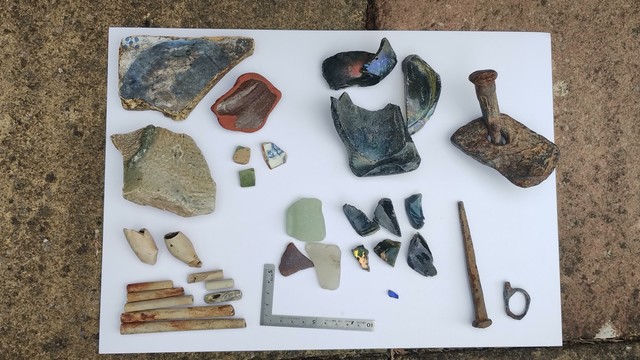 a display of mudlark finds from the Thames Foreshore. There is a partial blue and white tile, some other pottery, some broken clay pipes, broken thick old bottles and some ironmongery in the form of a broken scissors handle, a bit of rivet through metal plate and an old, long nail. There is a metal measuring square at the bottom for scale.