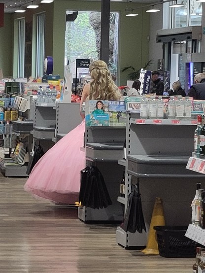a white lady, with a silver tiara in her blonde hair, wearing a pink ball gown is standing in line waiting to pay at a supermarket till.