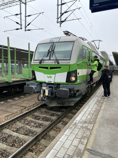 Kuljettaja kiipeää asemalaiturilta sähköveturin ovelle. Toinen mies seuraa vierestä. Veturin perässä on kaksikerrosmatkustajavaunuja, viereisellä raiteella tyhjiä puutavaravaunuja.