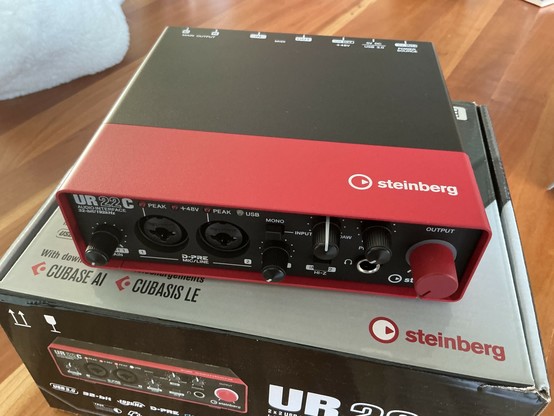 Image features a Steinberg UR22C audio interface, predominantly red with black accents. It includes two XLR inputs, various control knobs, and a USB connection. The device sits on top of its packaging on a wooden surface.