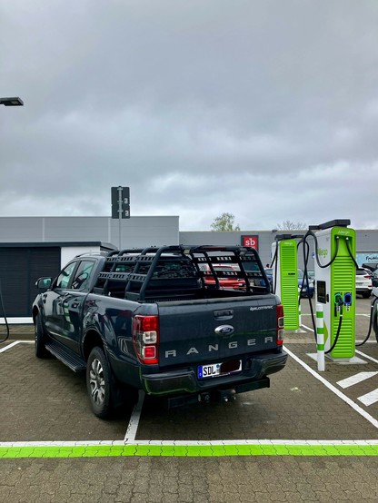 Ein dunkelblauer Ford Ranger Pickup ist in einem Laderaum neben einer grünen Ladestation für Elektrofahrzeuge geparkt. Der Himmel ist bedeckt, und es gibt umliegende Gebäude im Hintergrund.