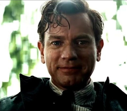 Ewan McGregor with longish top hair damp falling down onto his forehead. He's smiling, wearing a striped shirt, a dark tie, and overcoat. There are tree branches & leaves behind him. He is smiling and looking directly at the camera.