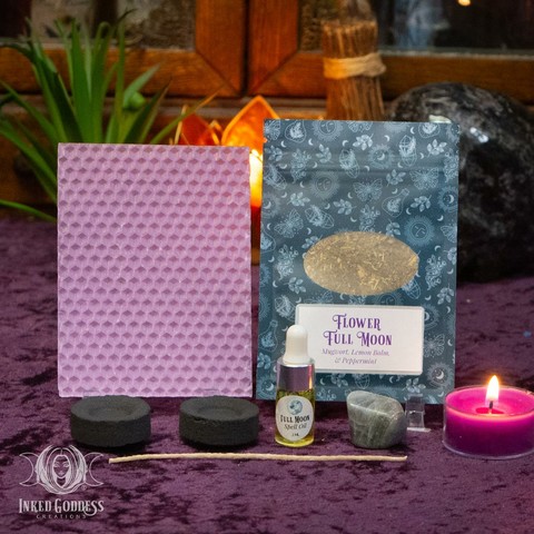 The contents of our exclusive "Flower Full Moon Ritual Kit for the May 12, 2025, Full Moon in Scorpio" product are arranged on a velvet-covered surface. They include a sheet of purple beeswax and a wick; a pouch of Flower Full Moon herbs, including mugwort, lemon balm, and peppermint; a dropper vial of our exclusive Full Moon Spell Oil; a tumbled labradorite gemstone; and two charcoal discs. Presented by Inked Goddess Creations.