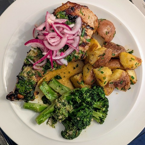 White plate, large, fat, char-marked pork chop topped with herb butter and red onions, skin-on potatoes with a scattering of chives, broccoli