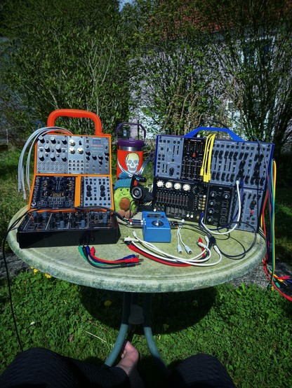 A starbase deep in Federation territory… captain’s log, stardate unknown. Two boldly colored, 3D-printed Eurorack cases (radiant orange and stellar blue) rest on a green, weathered garden table. Surrounded by blooming spring flora, the setup is ready for sonic warp trials. An orange thermos bottle adorned with a skull in Nonbinary pride colors stands tall beside the modules… radiating hydration and identity. The loyal officer AvocaJoe (knitted beanie included) guards the bridge. Multicolored patch cables arc like EPS conduits… connecting systems including a Stylophone drone synth below. Bare feet at the base… clearly, the Captain is grounded. Test phase initiated… tweaks may follow.