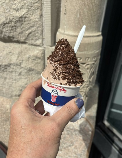 Paper “Mister Softee” cup of chocolate soft-serve, with chocolate crunch coating, a spoon, dripping over the edge. Background is a stone wall with engraving.