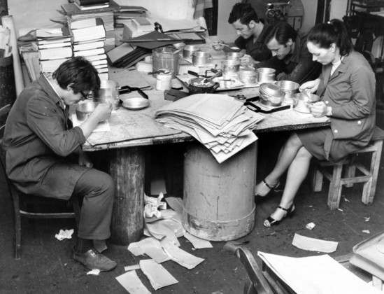 A black and white photo of four people in a factory setting eating lunch from metal pans at a large table that is covered in book and sheets of paper. The four people eating lunch are hunched over their food, concentrating hard and not appearing to talk to each other. The floor is also littered with various bits of paper.