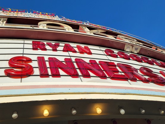 Marquee for movie Sinners, under a blue afternoon sky. The marquee is white with red block letters reading "Ryan Coogler's Sinners"