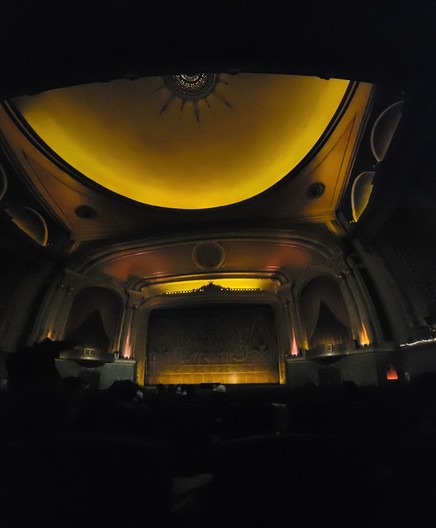 Panorama photo of the interior of the Gram Lake Theatre from right center seating. The lighting is pre-show, and yellowish. There is a dome at top with a decorative center with painted rays and stars. The main screen is covered by a sequined chain showing a scene of cornucopias, etc. Two box seats are at either edge of the stage. The feeling is very baroque and gilded.