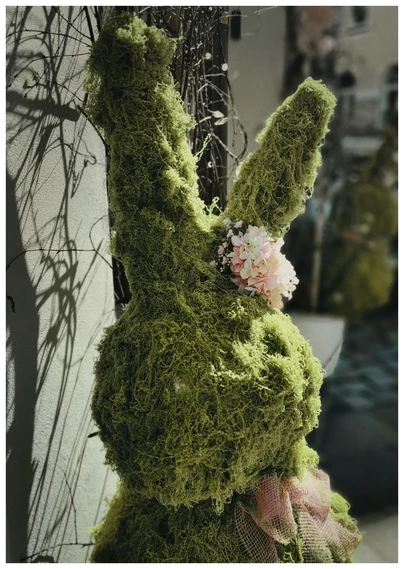 Formatfüllende Frontal-Aufnahme einer Deko-Hasenfigur aus hellgrünem, moosähnlichen Material. Die übergroße , gut 1,60 m hohe Dekofigur mit riesenlangen Löffelohren ist an einem Ohr am Ohransatz mit einer großen rosafarbenen Blume geschmückt, den Hals ziert eine gleichfarbene große Schleife. Die Figur stand - mit einem Zwilling - rechts und links am Eingang eines Ladenlokals auf einer belebten Geschäftsstrasse, die im Hintergrund unscharf zu erkennen ist.