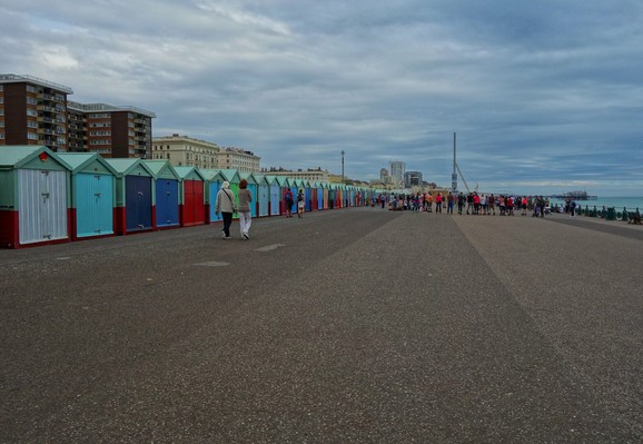 A wide shot captures a row of colourful beach huts lining the seafront. The huts are painted in various shades of blue, green, red and white. Two people are walking along the promenade in the foreground. In the background, buildings and a pier stretch into the distance under an overcast sky. A group of people are gathered further down the promenade.