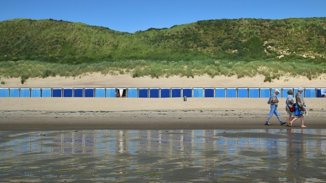 ein bild von drei älteren menschen, die am strand entlanggehen, vor einer reihe blauer strandkabinen. im hintergrund befinden sich hohe sanddünen mit grüner vegetation. das wasser des meeres spiegelt die strandkabinen wider. der himmel ist blau und wolkenlos. es ist ein sonniger tag. die szene strahlt ruhe und entspannung aus. es könnte ein typischer sommertag an der küste sein.