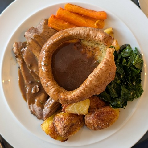 White plate, slices of roast lamb, carrots, parsnips just peeking out, greens, roast potatoes and a big herby pudding, gravy sluiced over