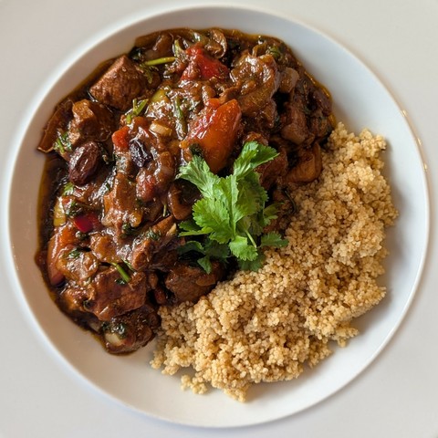 White bowl, large mass of dark brown stew chunky with lamb, peppers, olives and fruit, pile of fluffy couscous