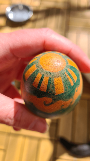 Photo. Egg is angled to show more detail of the sun drawing. A circle with squared-off rays coming from it. Held in hand.