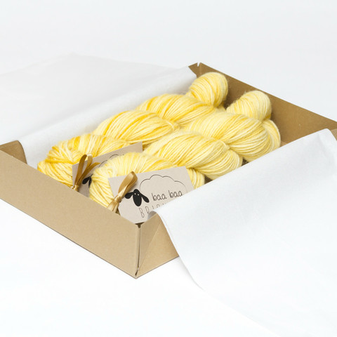 Picture shows an open A4 box inlaid with white tissue paper containing two skeins of yarn in shades of yellow.
