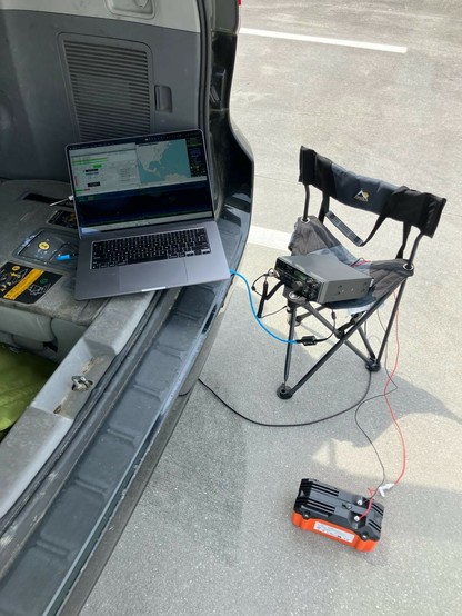 A laptop, gray transceiver, and orange battery arranged around the open back of a minivan.