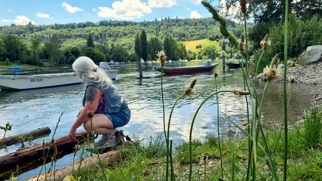 Rheinufer, Kähne, Holzbohlen im Wasser, hügelige Landschaft, blauer Himmel, glitzerndes Wasser in allen blau-grün-Schattierungen, eine Frau in der Hocke mit langen weißen Locken und in kurzer Jeans und Shirt