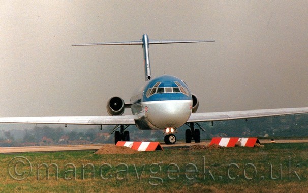 Almost head-on view from slightly below of a blue and grey, twin engined jet airliner with the engines mounted on the sides of the rear fuselage, turning to it's right as it lines up on a runway.
The upper body is a dark blue, with a grey belly, a white stripe running along the body separating the 2 sections.
A small British flag is visible on each side of the lower forward fuselage, under the rear cockpit windows.
Green grass fills the foreground, with piles of dirt on the ground, with red and white striped triangular frames making their location.
Under the planes wings, trees and houses can be seen in the distance, with dismal grey sky filling the rest of the frame.