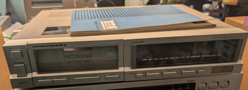 A strange looking CD player on a workbench with a manual on top. The left side is dominated by a window showing three leds and some keys (rev, fw, next program, pause). On the right, there is another big window with a row of lightbulbs and another row of LEDs beneath them. Underneath, another set of buttons ("Stop / All cancel", "Select", "Preset", "Repeat", "Cancel")