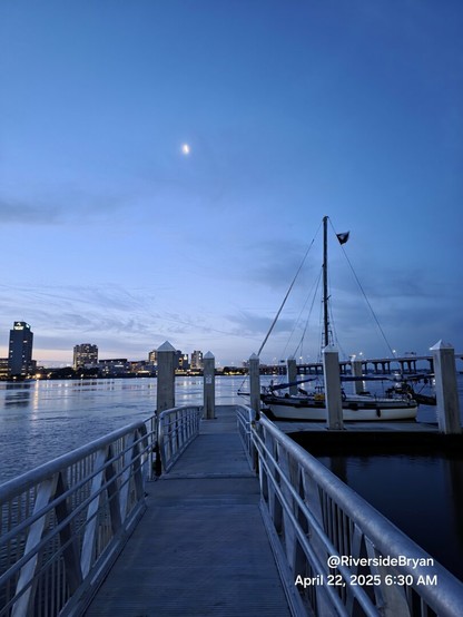 Early morning deep blue sky. Illuminated by a crescent moon above a boat dock gangway leading to a sailboat tied up on the river.