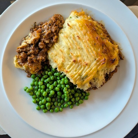 White plate, slab of shepherd's pie with crusty fork-marked mash top and a little spill of lamb mince interior, pile of peas