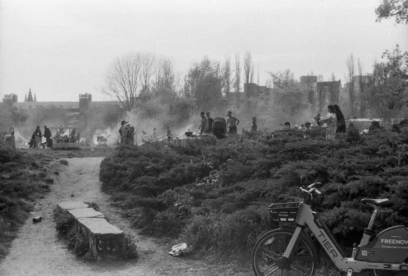 ein schwarzweißes photo aus dem mauerpark, im vordergrund ein leihfahrrad von TIER, im hintergrund allerlei parkgäste an grills und viel rauch, weiter hinten häuser